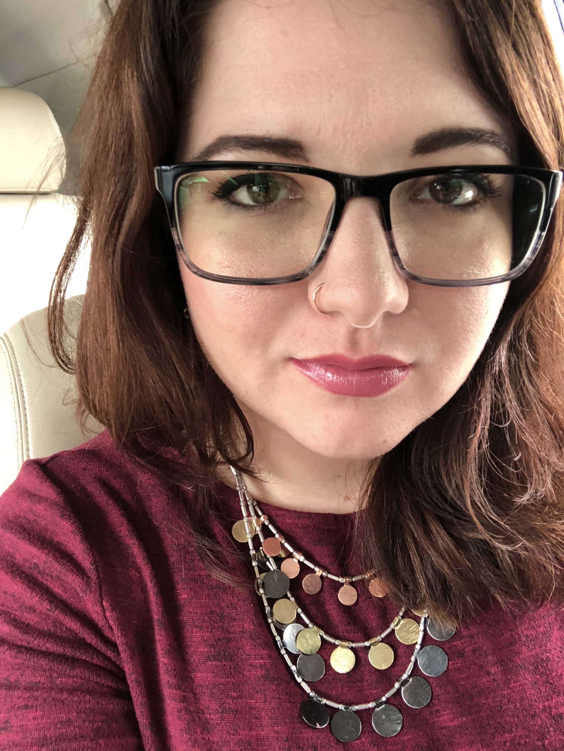 Close up image of Jessica Lee Gray, an adult woman with auburn hair wearing a red sweater, with multilayered coin necklace and black framed glasses.