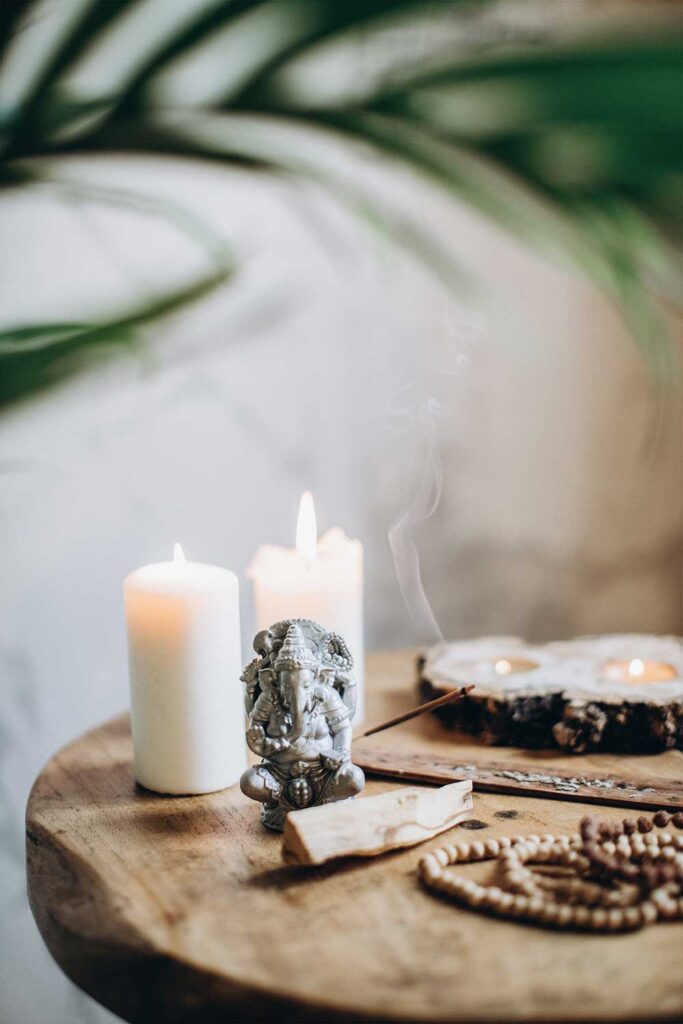 Decorative image, foreground has shallow depth of field featuring a rustic, circular wooden table, two white pillar candles, palo santo stick, Ganesh sculpture, wooden prayer beads and burning incense. There is a palm frond draped across the white background.
